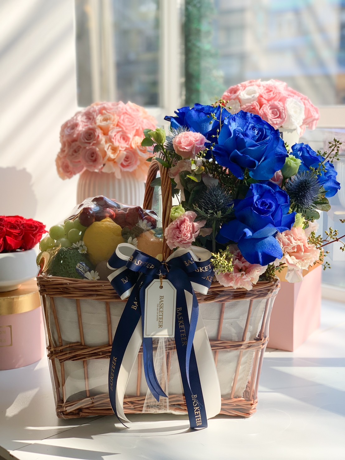 A luxurious fruit and floral basket featuring royal blue roses, pink flowers, and premium fresh fruits, beautifully arranged in a woven basket with a navy blue and gold Basketeer ribbon.