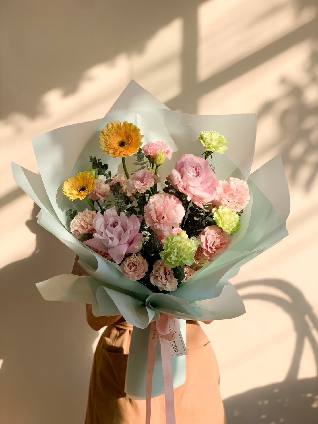 A charming pastel bouquet with pink roses, peach carnations, yellow gerberas, and greenery, wrapped in soft mint green with a Basketeer ribbon.