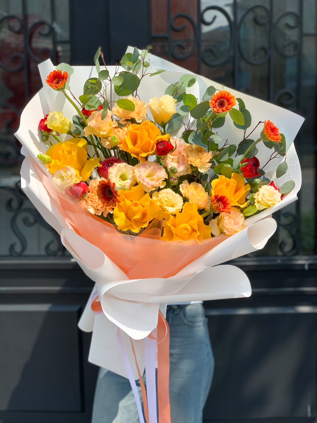 A vibrant Sunset Bloom Bouquet featuring yellow roses, peach lisianthus, orange gerberas, and red ranunculus, elegantly wrapped in soft white and peach paper.