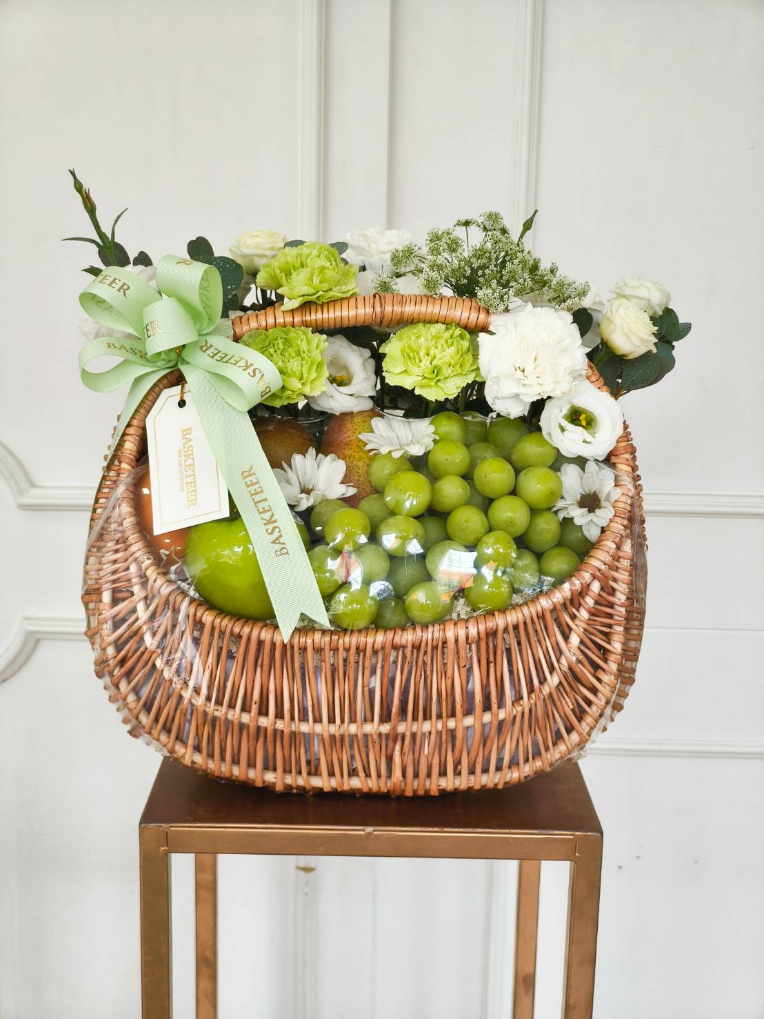 Luxury wicker fruit basket filled with fresh green grapes, pears, and apples, adorned with green and white flowers and a green satin bow.