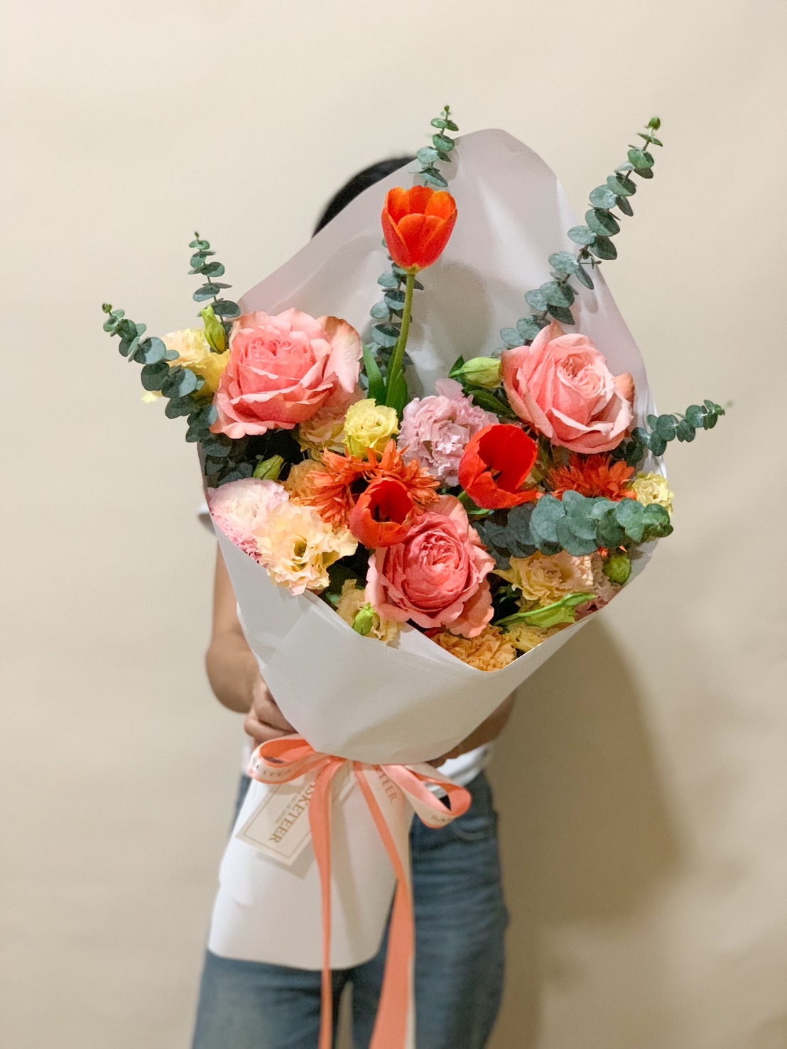 A stunning mixed flower bouquet with roses, tulips, eucalyptus, and lisianthus, wrapped in elegant paper with a peach ribbon.