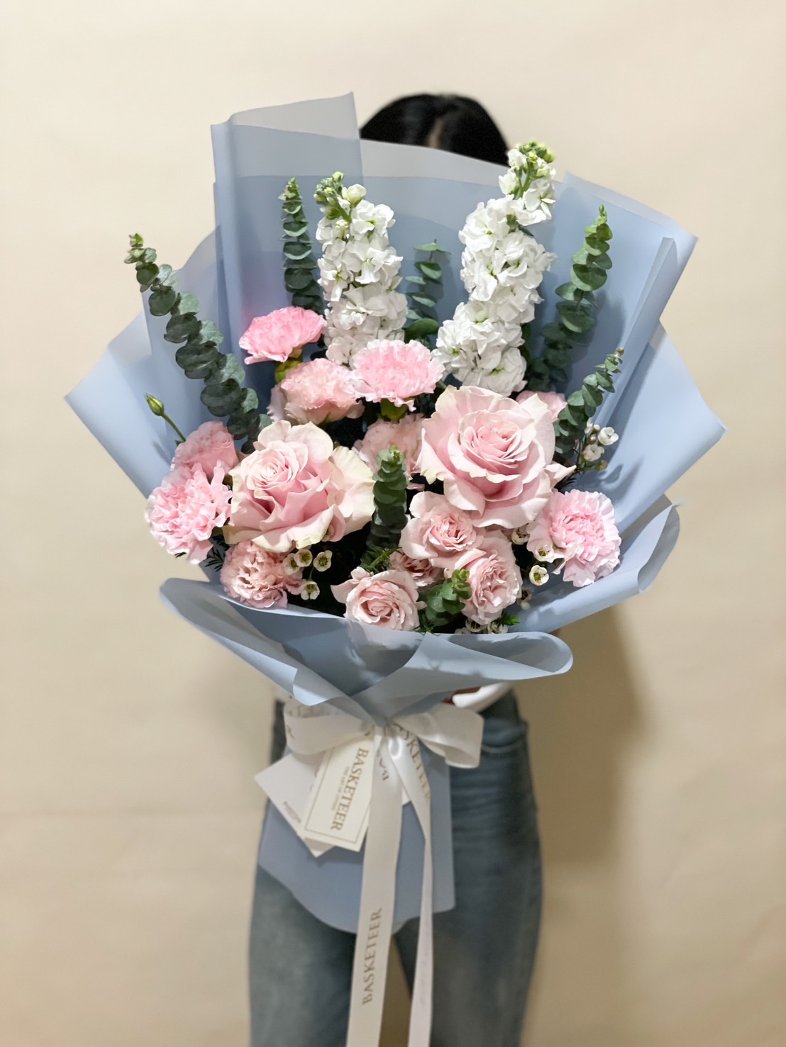 A beautifully arranged pastel pink and white bouquet, featuring roses, carnations, and stock flowers, wrapped in soft blue paper with a white ribbon.
