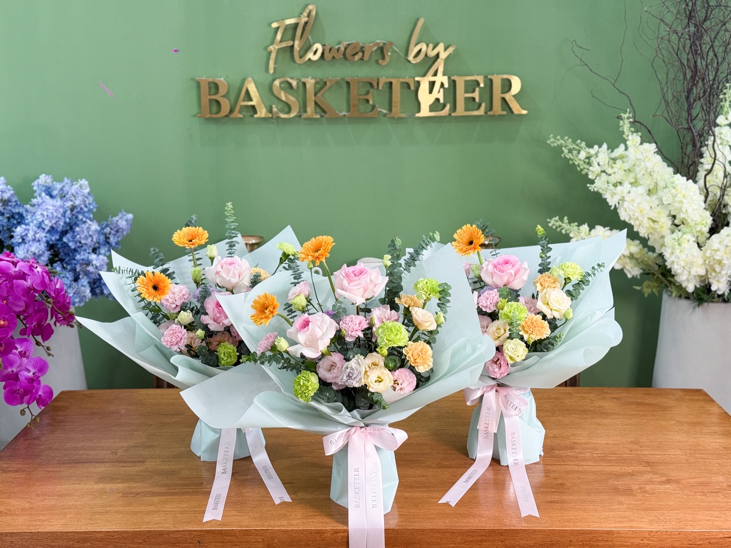 Elegant pastel flower bouquets featuring pink roses, peach carnations, yellow gerberas, and green blooms, wrapped in mint paper with white ribbons.