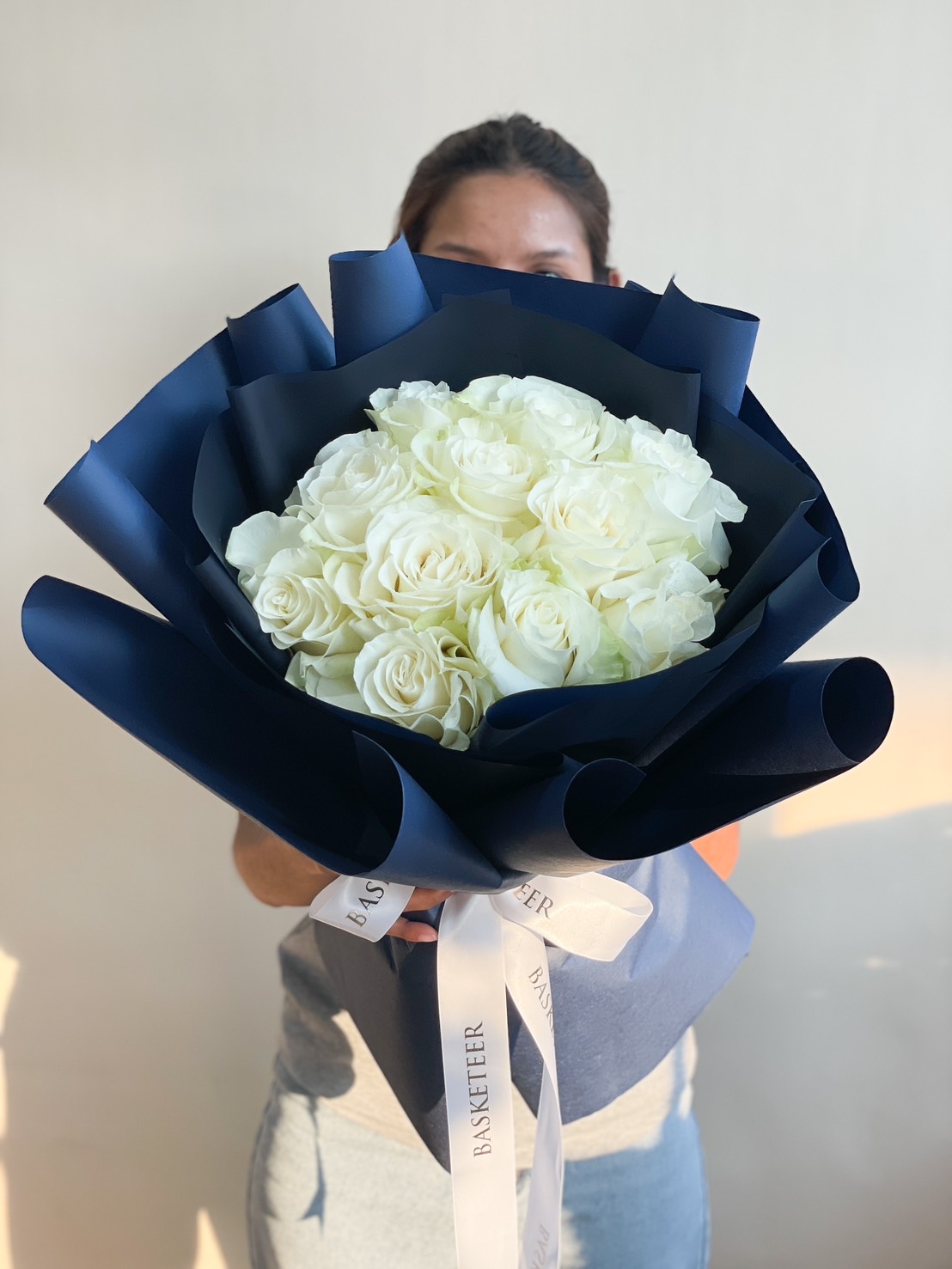 A lavish bouquet of fresh white roses, wrapped in elegant deep blue paper with a white Basketeer ribbon, symbolizing pure love and timeless beauty.