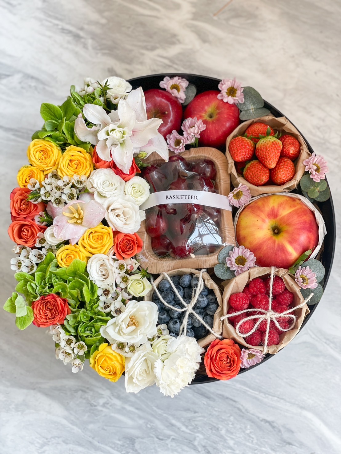 A luxurious fresh fruit gift basket featuring apples, strawberries, raspberries, blueberries, and cherries, elegantly arranged with roses and delicate blooms in a premium black round box.