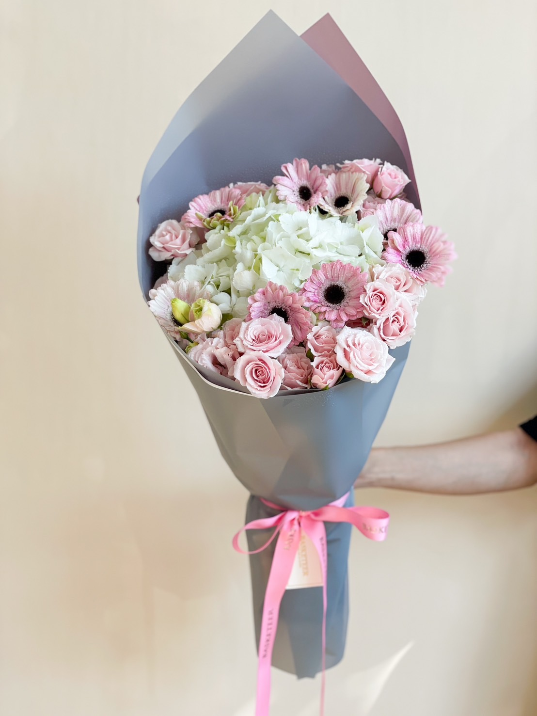 A sophisticated pink and white floral bouquet featuring white hydrangeas, pink gerbera daisies, and blush roses, wrapped in stylish dark-toned paper with a pink ribbon.