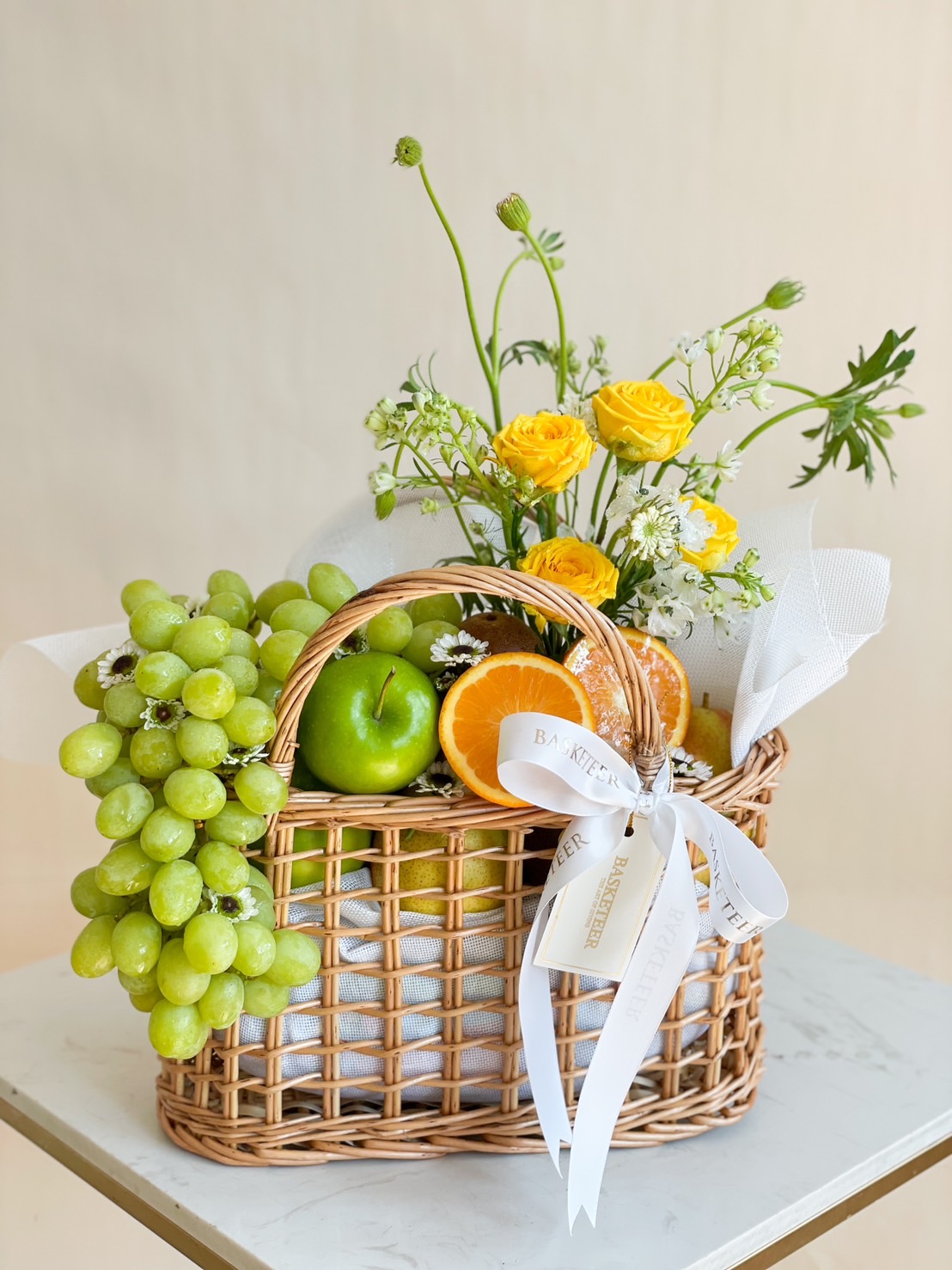 A premium fruit & flower gift basket filled with green grapes, apples, oranges, and vibrant yellow roses, elegantly arranged in a woven wicker basket with a white ribbon.