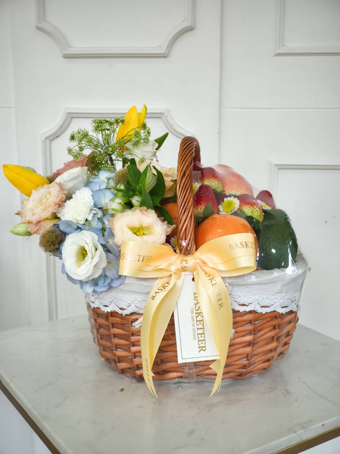 Premium wicker fruit and flower basket with oranges, apples, avocado, yellow lilies, blue hydrangeas, and lisianthus.