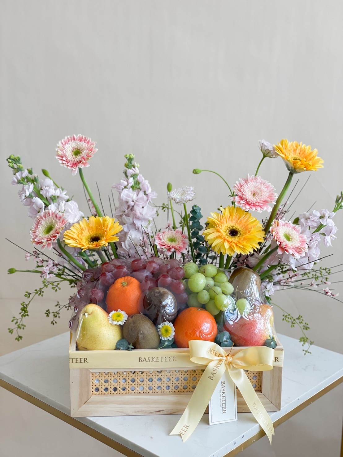 A beautifully arranged luxury fruit and flower gift basket, featuring fresh grapes, oranges, pears, kiwis, and apples, surrounded by vibrant yellow, pink, and purple flowers in an elegant wooden basket with a golden ribbon.