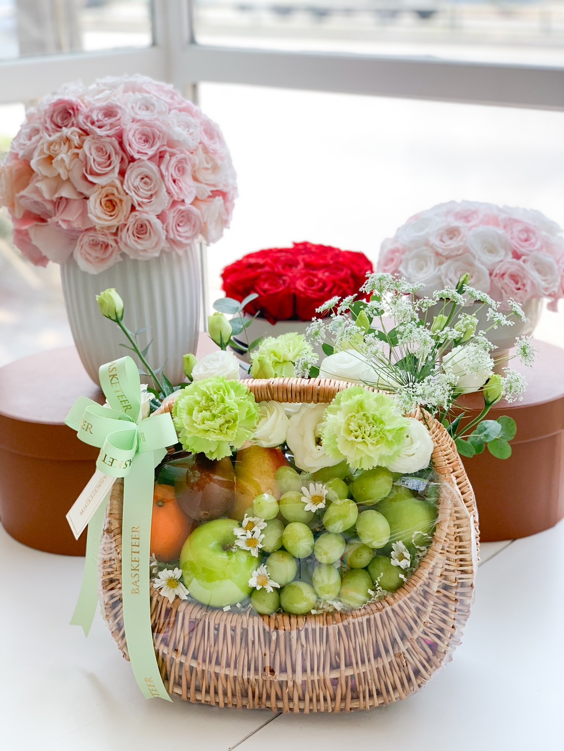 A beautifully arranged green-themed fruit basket filled with fresh green apples, grapes, pears, and citrus fruits, decorated with white and green carnations, delicate daisies, and wrapped in an elegant wicker basket with a matching pastel green ribbon.