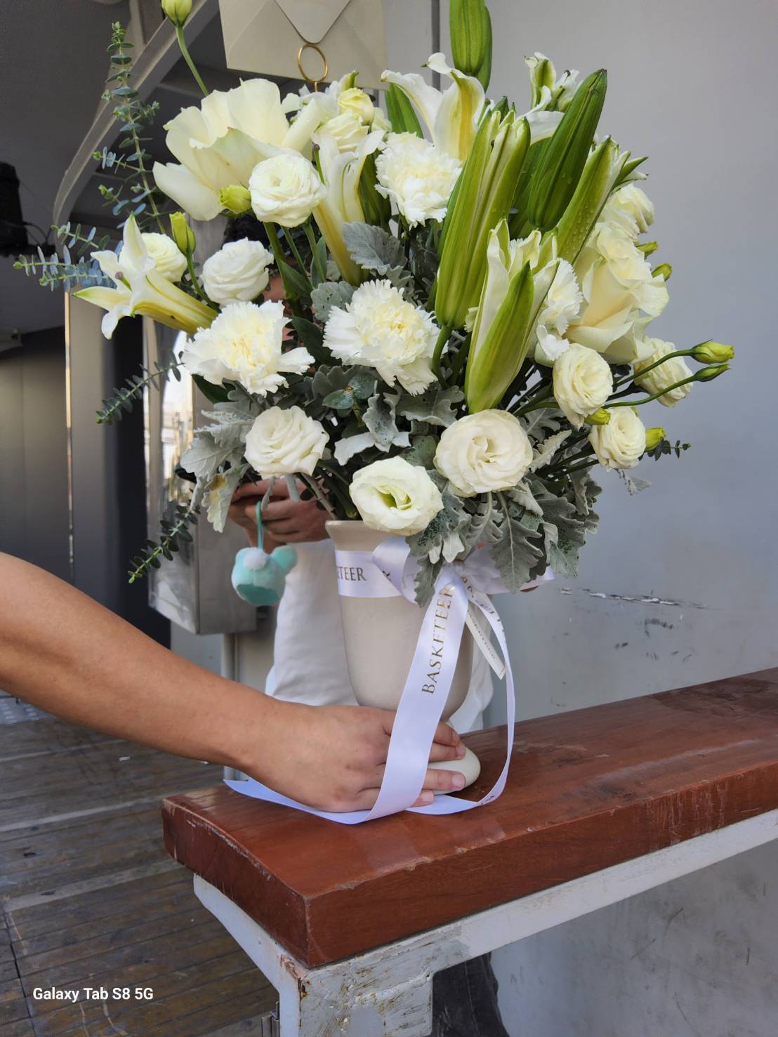 A stunning white floral arrangement with fresh white lilies, roses, and carnations elegantly displayed in a decorative vase, wrapped with a white satin ribbon.