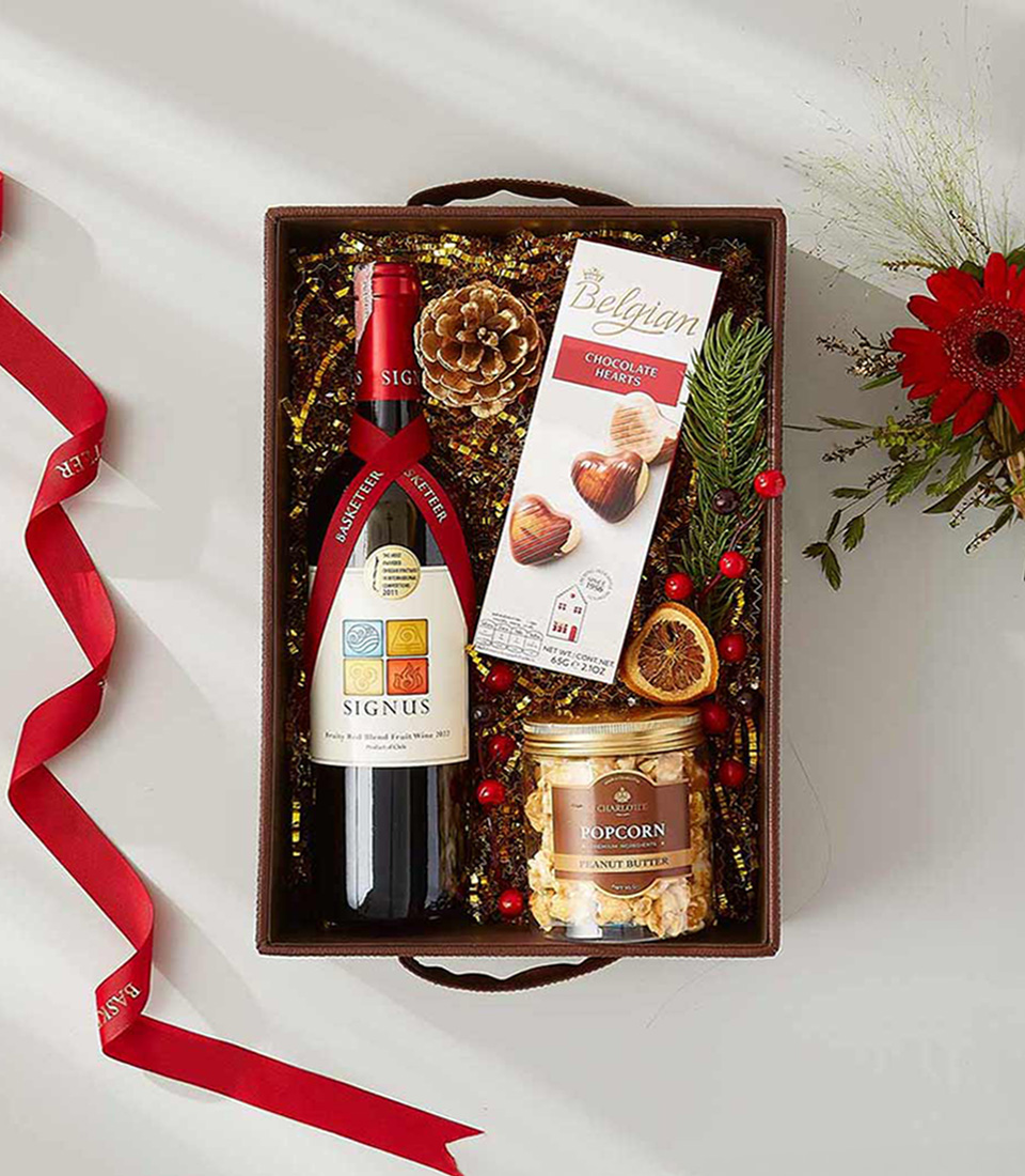 A brown gift box filled with Christmas goodies, including a bottle of red wine, a box of Belgian chocolate hearts, a jar of peanut butter popcorn, and festive decorations.