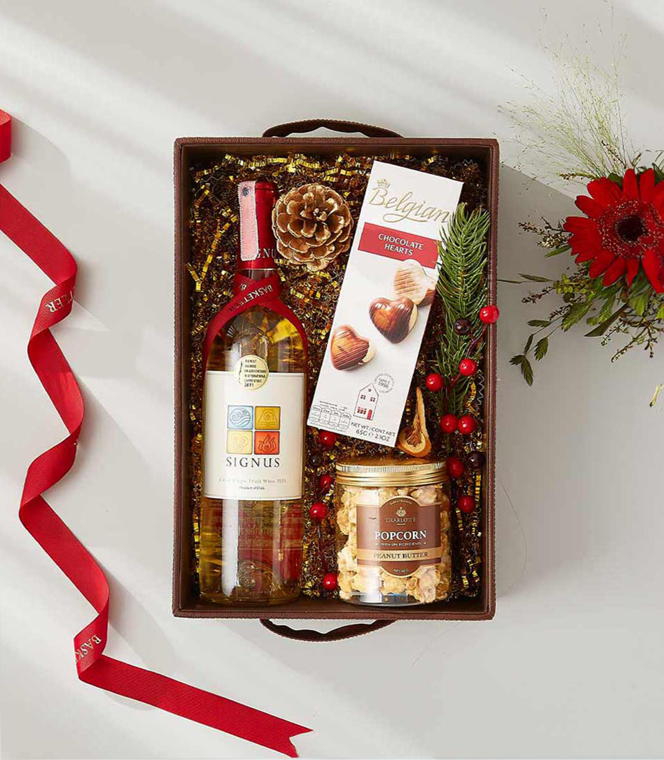 A festive gift basket overflowing with gourmet treats, including a bottle of white wine, Belgian chocolate hearts, and a jar of peanut butter popcorn.