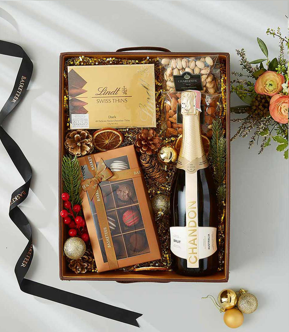 A festive Christmas gift box, adorned with a red satin ribbon, brimming with a bottle of Chandon Brut sparkling wine, an assortment of Lindt Swiss Thins, rich gourmet chocolates, and a selection of gourmet nuts.