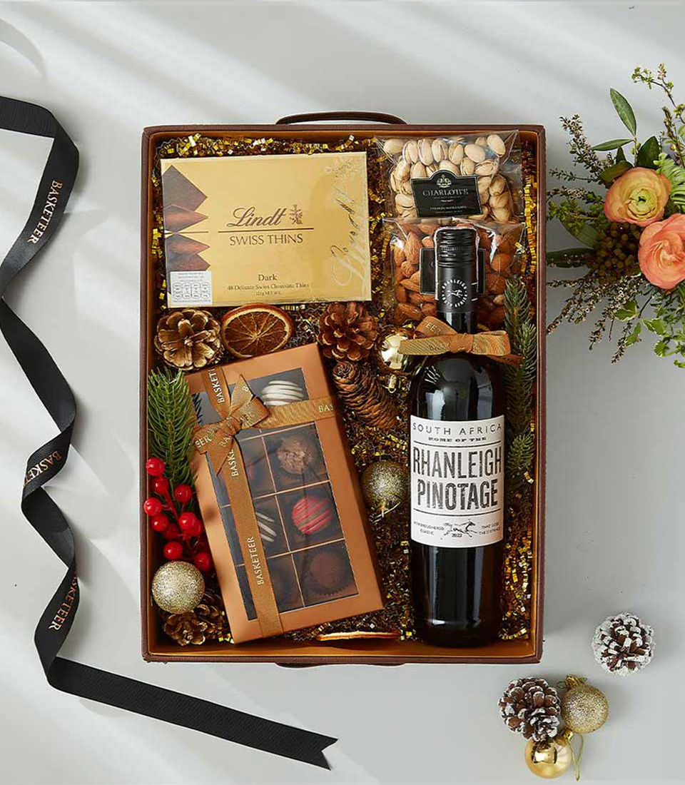 A festive Christmas gift box, adorned with a thick red satin ribbon, brimming with a bottle of South African Pinotage wine, a selection of rich gourmet chocolates