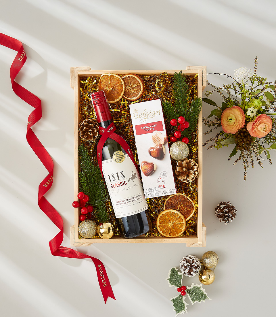 A wooden crate filled with holiday cheer, featuring a bottle of red wine, Belgian chocolate hearts, and festive decorations.