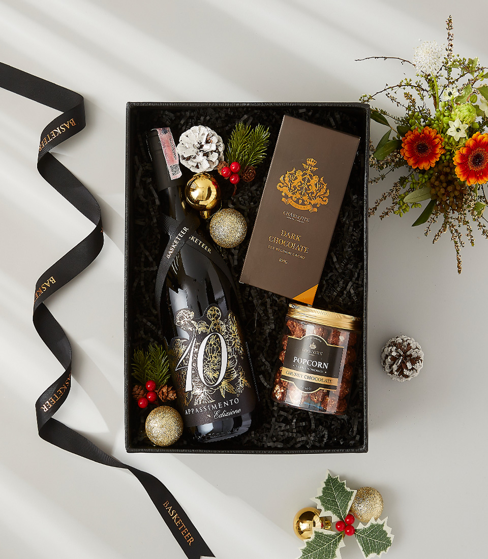 A black gift box filled with holiday cheer, featuring a bottle of wine, dark chocolate, popcorn, and festive decorations.