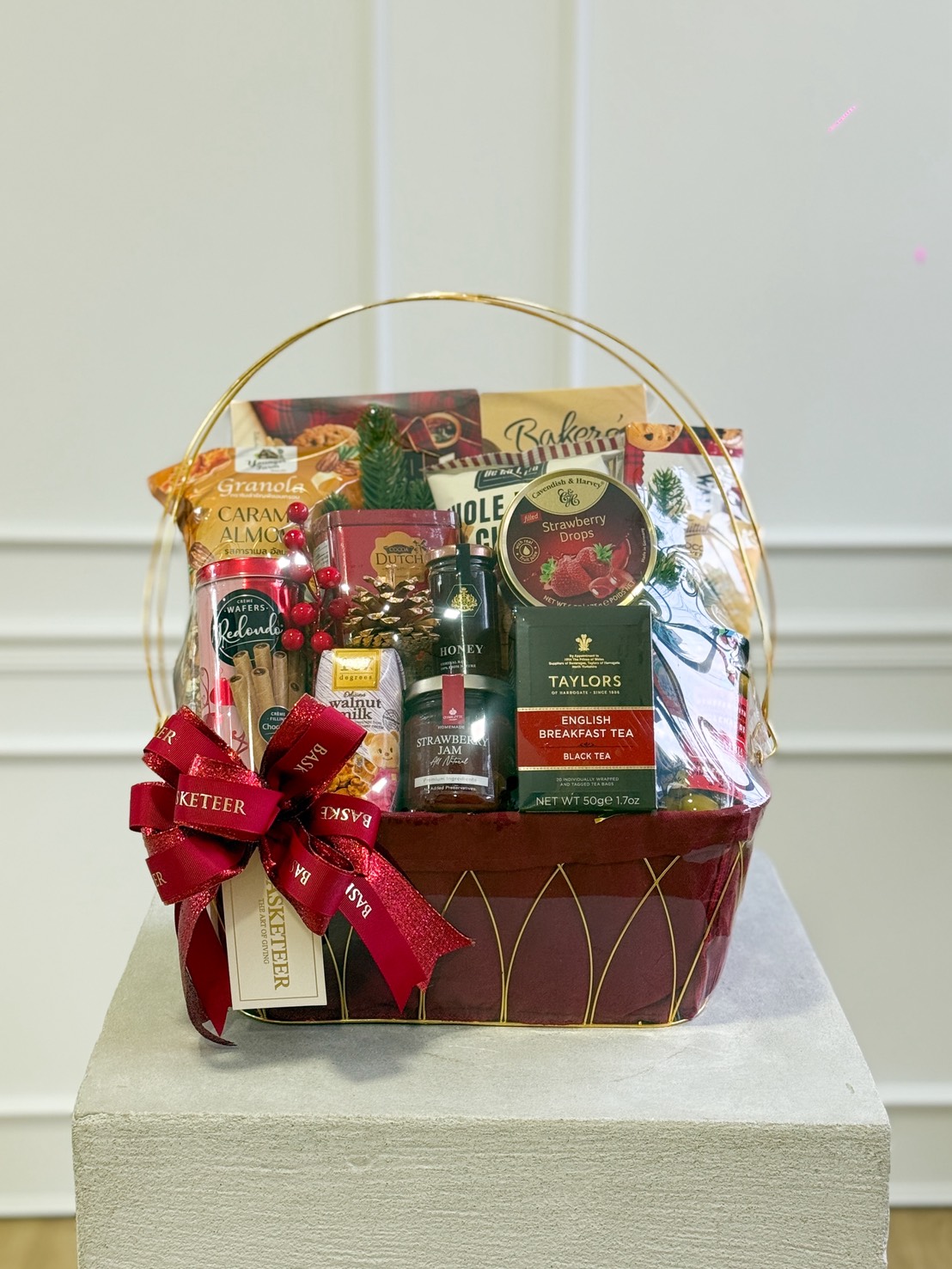 A Festive Treats Gift Hamper with assorted treats, tea, chocolates, biscuits, and snacks, wrapped in clear plastic and decorated with a red ribbon and greenery, displayed on a pedestal against a white wall.
