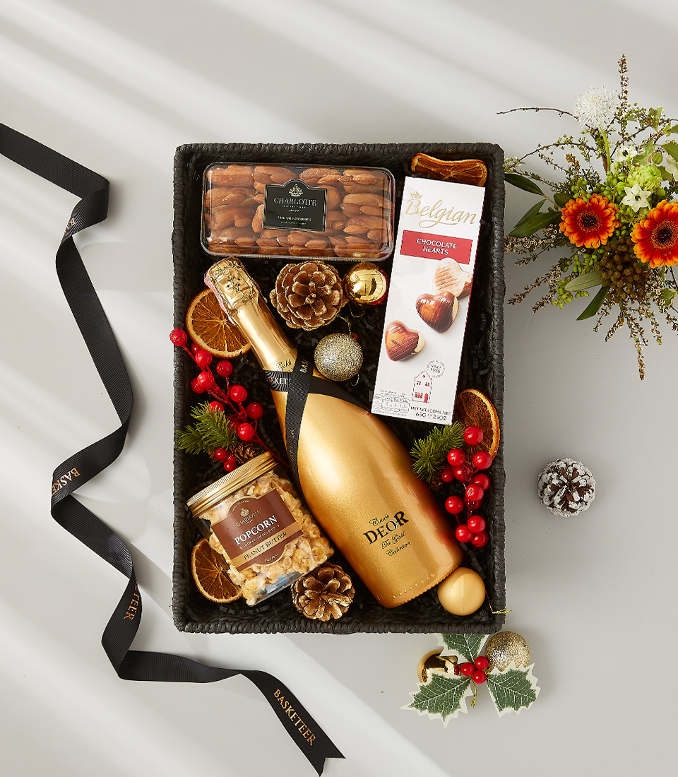 Festive gift basket containing a bottle of golden champagne, gourmet chocolate, popcorn, and almonds. Decorated with pine cones, dried oranges, and holly.