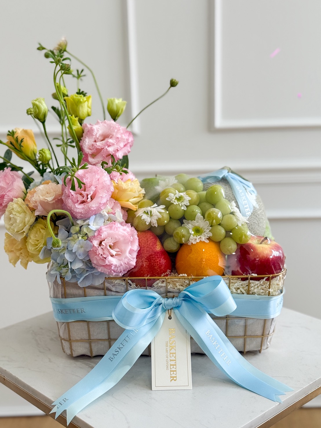 A Pastel Harmony Fruit Basket filled with green grapes, apples, oranges, and topped with pink and yellow flowers, decorated with light blue ribbons and a 