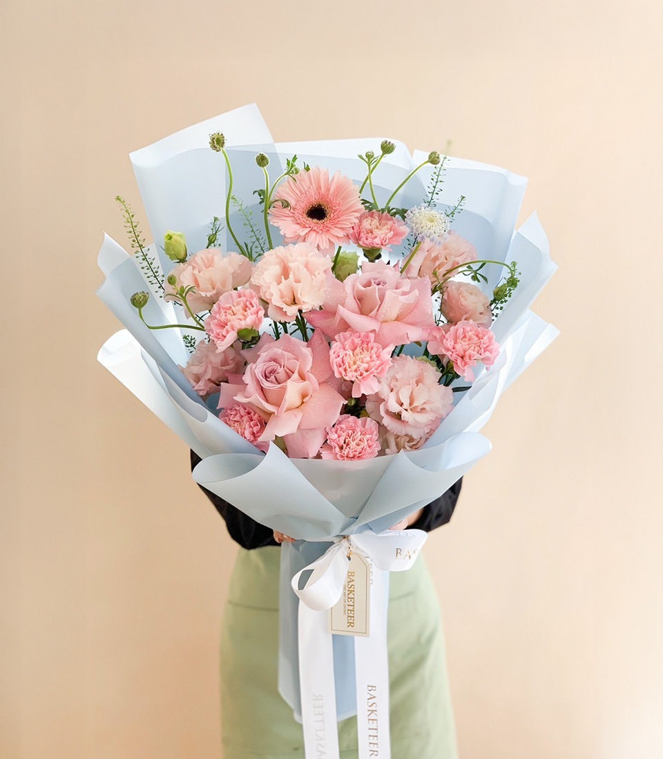 Soft pink bouquet with roses, carnations, and gerberas wrapped in pastel blue paper with a white ribbon.