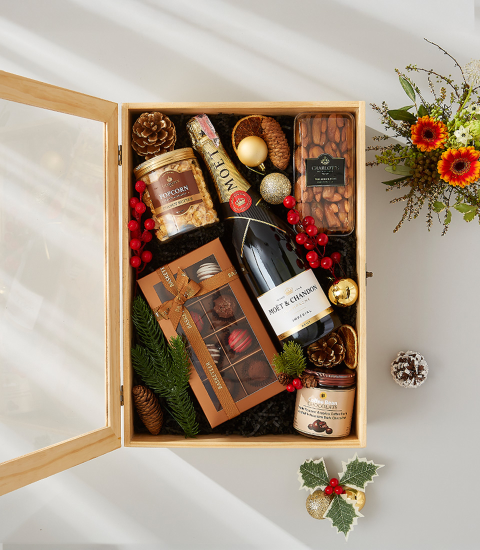 A wooden gift box filled with a bottle of Moët & Chandon champagne, a box of gourmet chocolate truffles, and a variety of festive snacks.