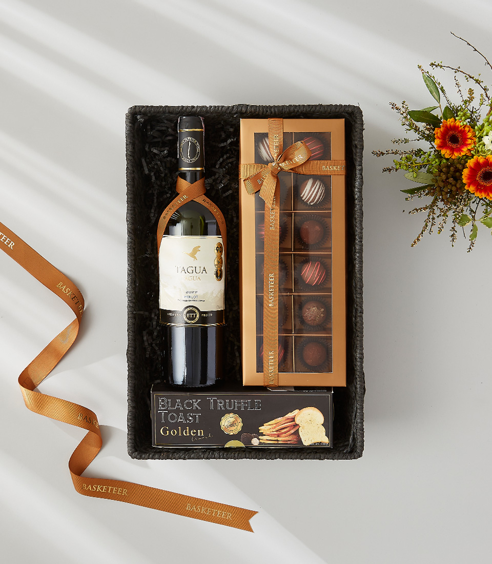 A rectangular gift basket filled with a bottle of wine, a box of chocolates, crackers, and decorative ribbon.