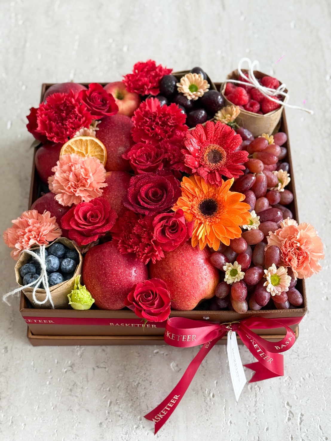A decorative fruit box arranged with red apples, grapes, blueberries, strawberries, orange slices, and assorted red and orange flowers—an exquisite Japanese Melon & Rose Gift Hamper—tied with a red ribbon on a light background.