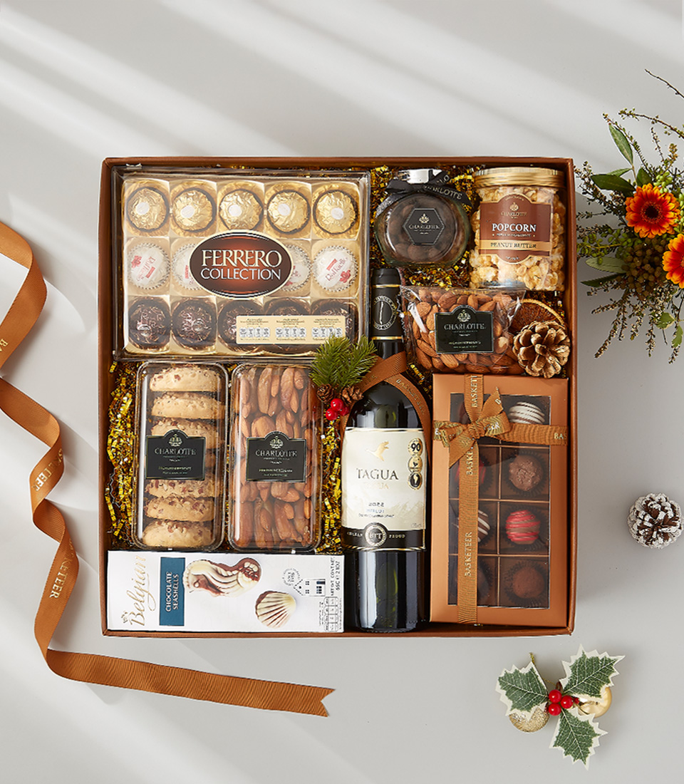 A rectangular gift box filled with various gourmet treats, including a bottle of red wine, a box of Ferrero Rocher chocolates, a jar of chocolate spread, cookies, nuts, popcorn, and holiday decorations.