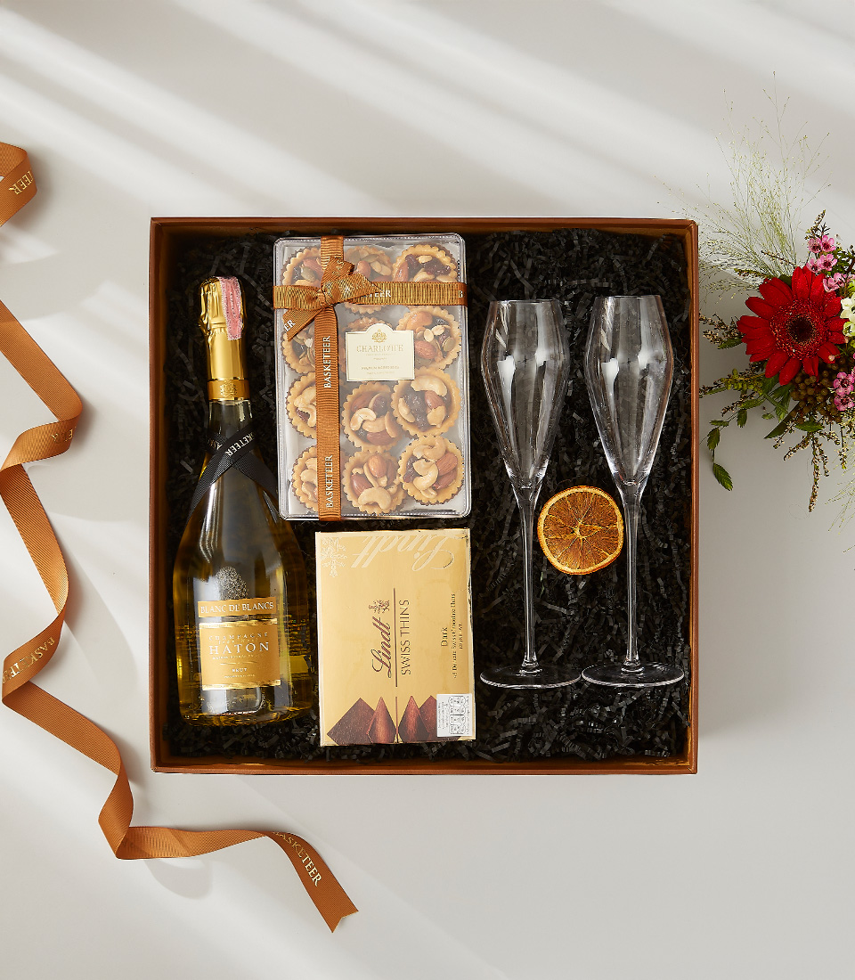 A gift box containing a bottle of champagne, Swiss chocolates, gourmet nut toasts, and two champagne flutes with a dried orange slice.