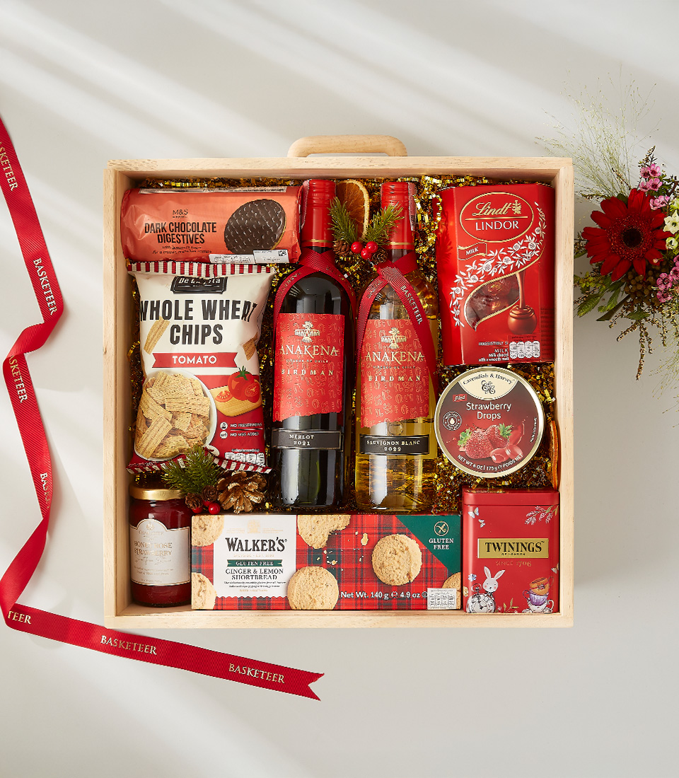A wooden crate filled with a variety of Christmas treats, including a bottle of red wine, cookies, nuts, chocolates, and popcorn