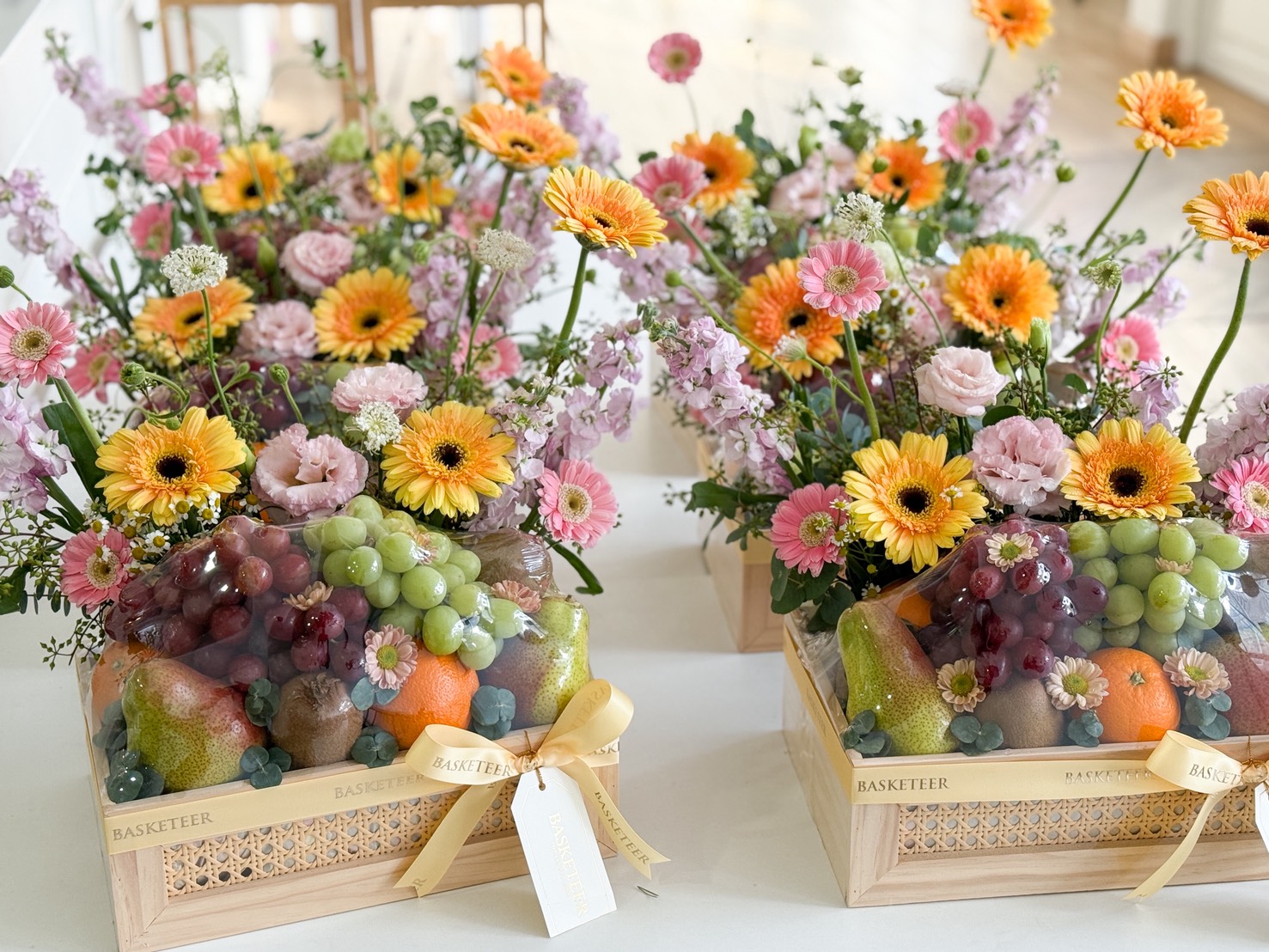 The Garden Bloom Fruit Box features wooden crates filled with colourful mixed flowers—yellow gerbera daisies, pink blooms—and an assortment of grapes, oranges, and pears, each crate adorned with a yellow ribbon and gift tag.