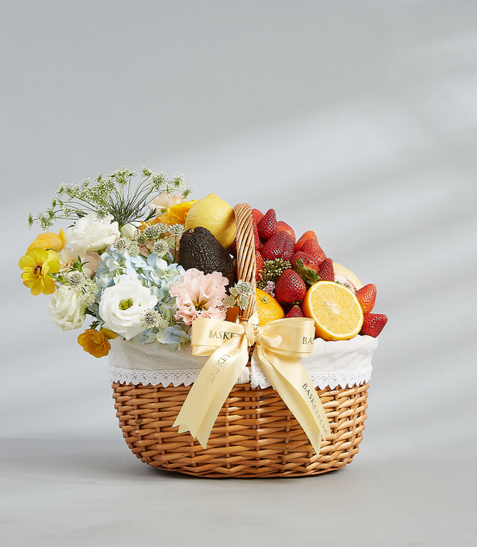 Elegant fruit and flower basket with fresh strawberries, lemons, avocados, oranges, and a bouquet of white, yellow, and pastel flowers.