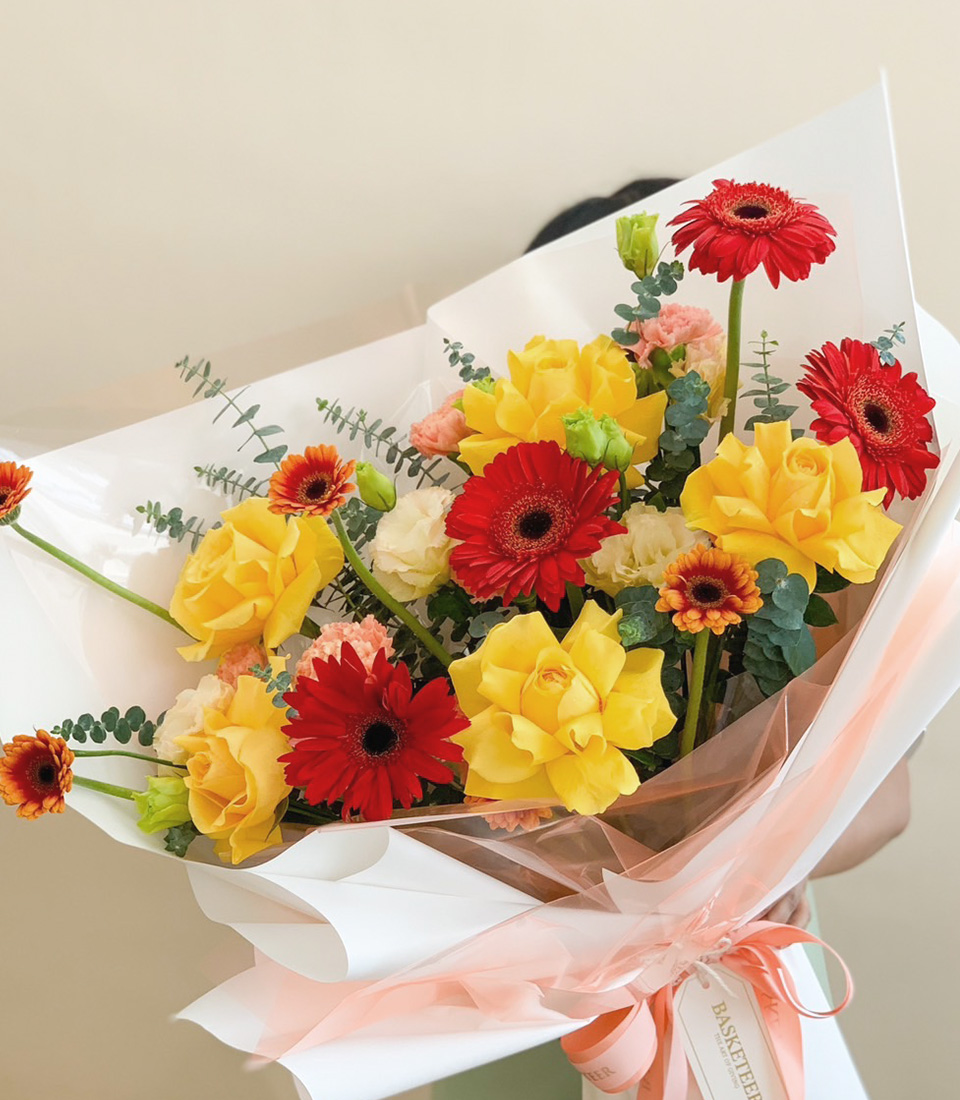 A vibrant bouquet of yellow roses and red gerberas with greenery, wrapped in soft white and peach paper, tied with a peach ribbon.