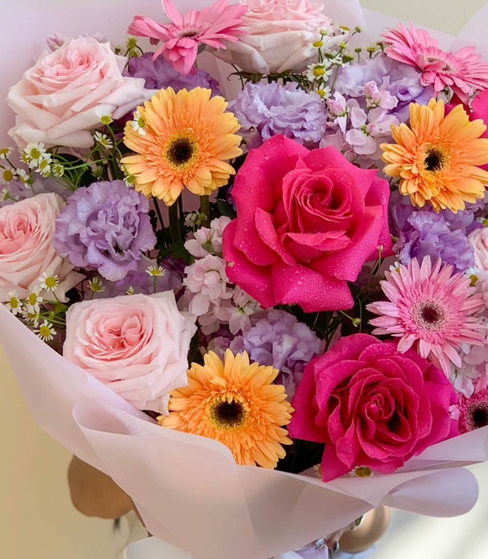 A large bouquet of pink roses, gerberas, purple lisianthus, and orange blooms, wrapped in soft pink paper and tied with elegant ribbons.