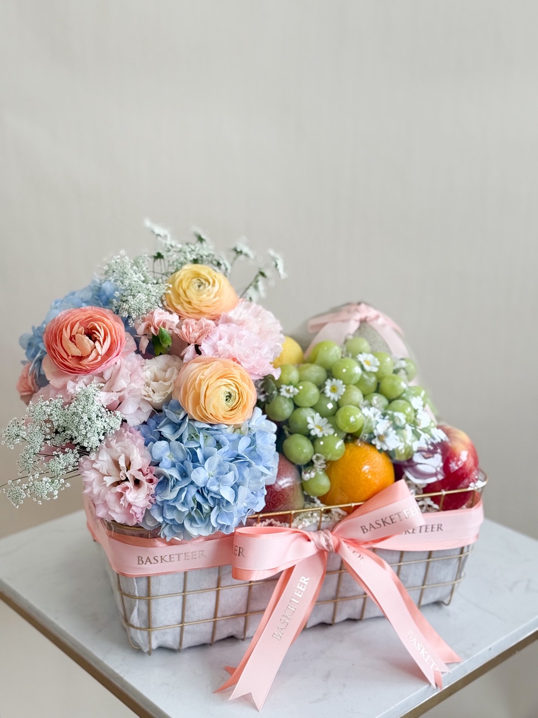 Premium fruit basket with pastel flower arrangement including hydrangeas, ranunculus, carnations, grapes, apples, and oranges.