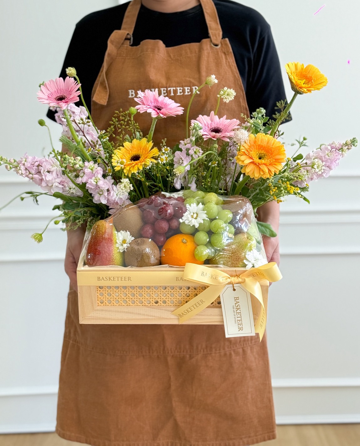 A person wearing a brown apron holds a wooden basket filled with assorted fresh fruit and colourful flowers, decorated with a cream ribbon, a 