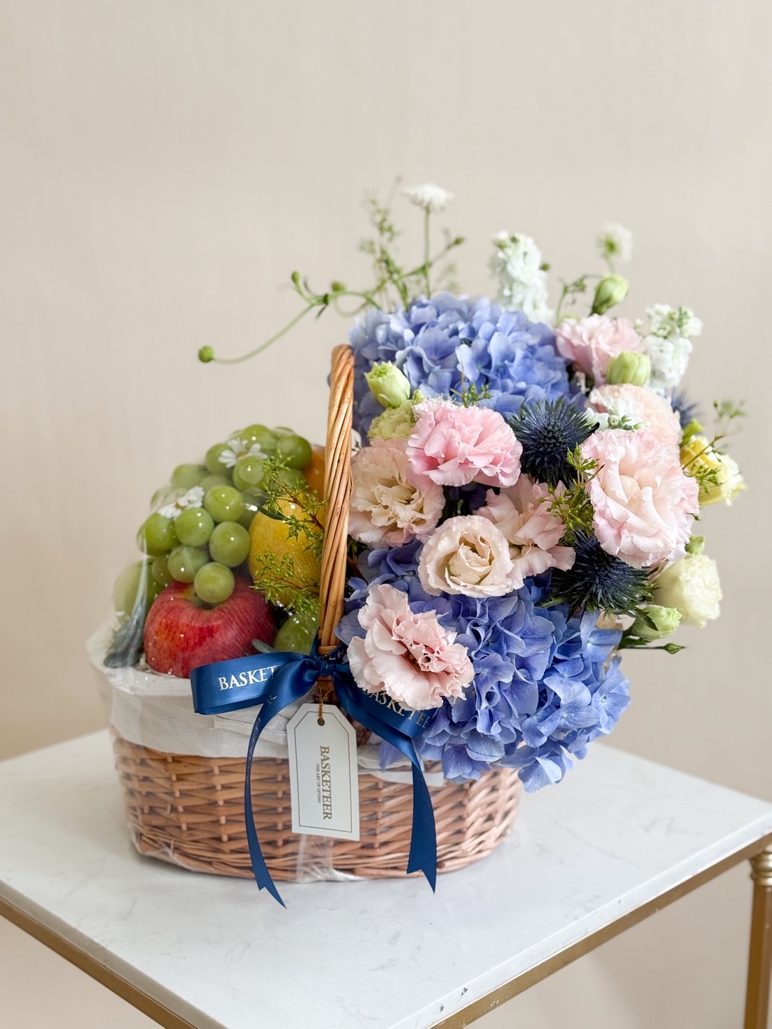 : Luxury fruit basket with green grapes, apples, and assorted pastel flowers including blue hydrangeas and pink lisianthus in a woven basket