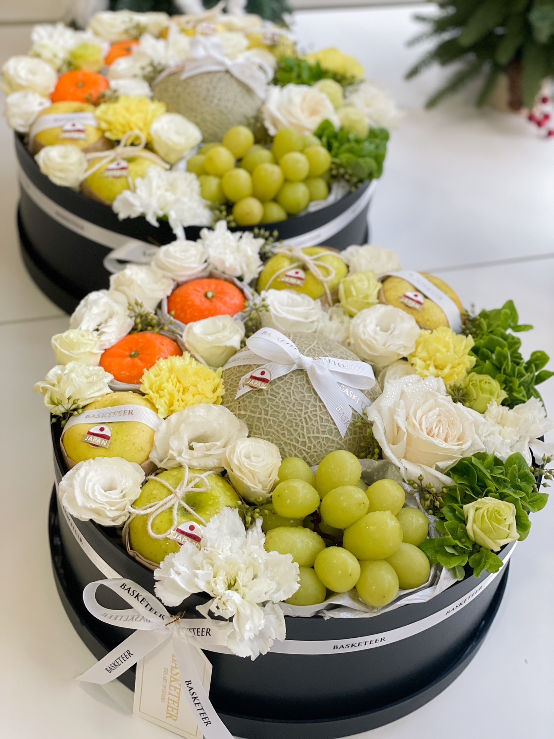 Elegant Japanese fruit gift box with fresh melon, green grapes, oranges, pears, and white seasonal flowers in a black round box.