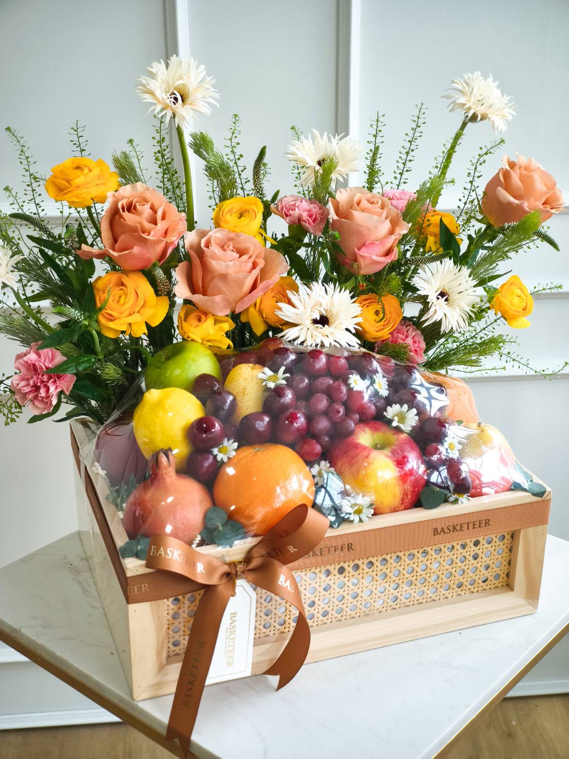 Luxury fruit and flower basket with apples, grapes, oranges, pomegranates, and roses in yellow and orange tones, tied with brown ribbon