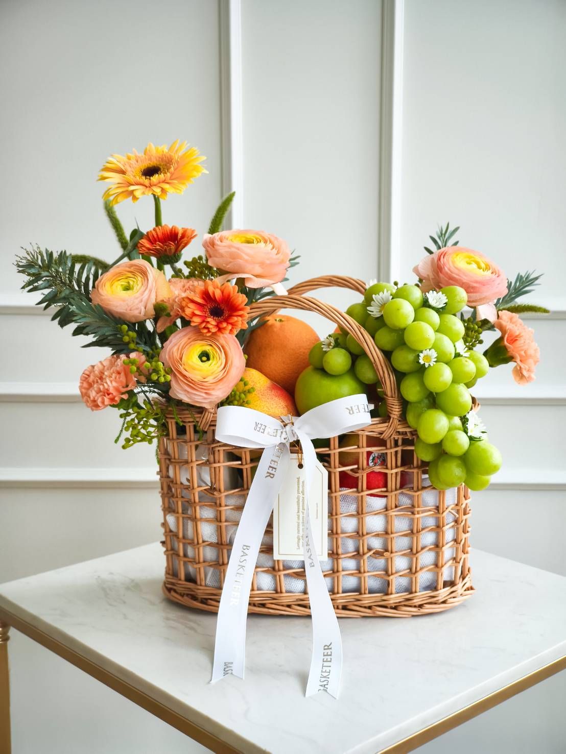 Premium fruit basket with green grapes, oranges, apples, and orange-yellow flowers in a woven basket with ribbon