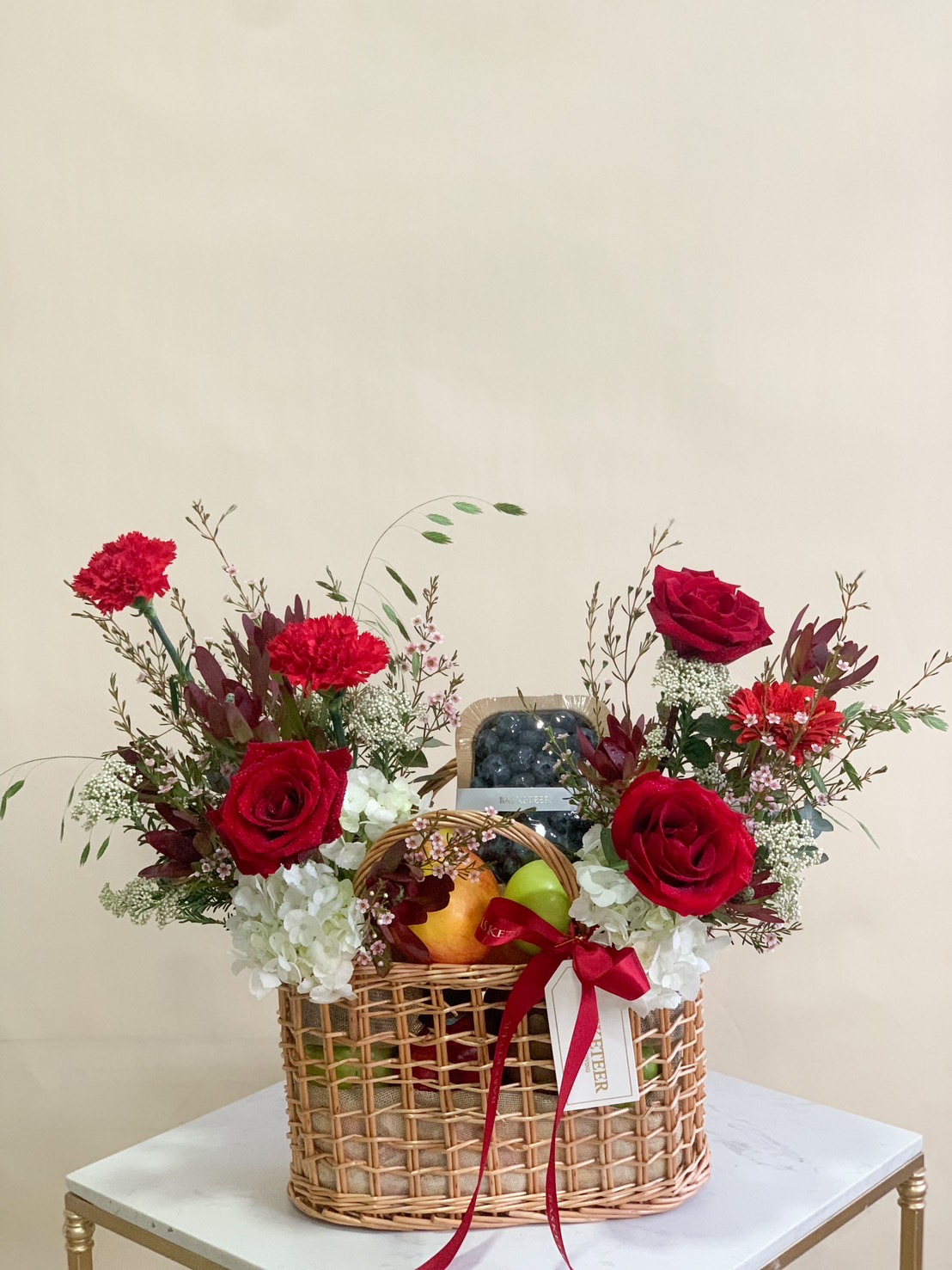 Luxury fruit and flower basket with red roses, carnations, white hydrangeas, and assorted fresh fruits in a woven basket.