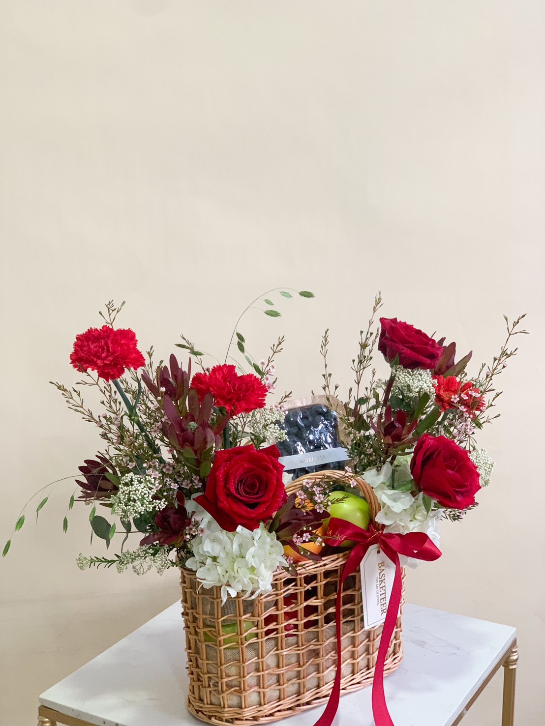 Luxury fruit and flower basket with red roses, carnations, white hydrangeas, and assorted fresh fruits in a woven basket.