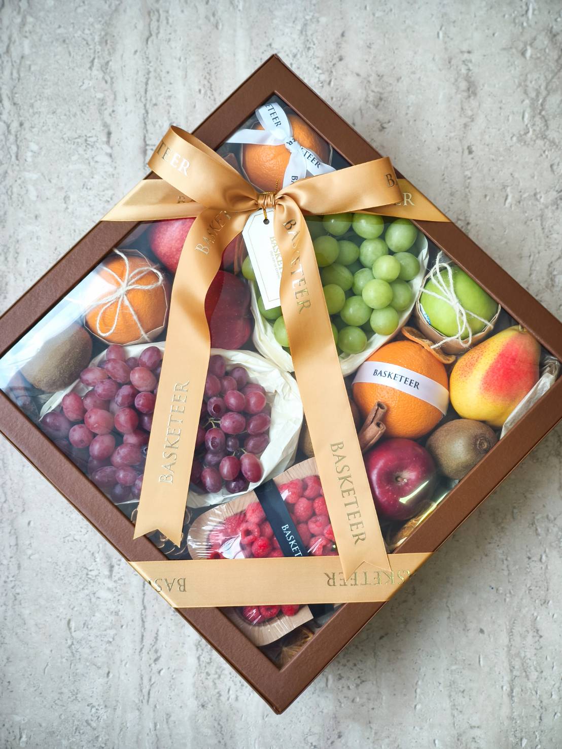Premium fruit gift box with green grapes, red grapes, apples, pears, oranges, kiwis, and raspberries, tied with a gold satin ribbon