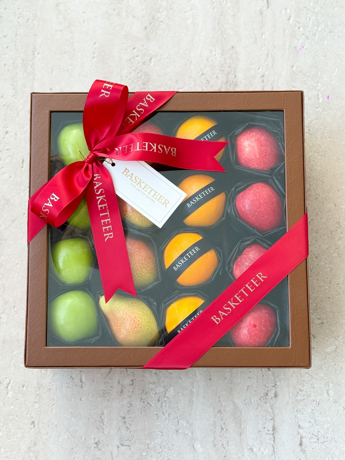 A Deluxe Fruit Gift Box, this brown box with a clear lid displays assorted artificial fruits—apples, oranges, and pears. Finished with a red "Basketeer" ribbon and matching tag, it makes for an elegant presentation.