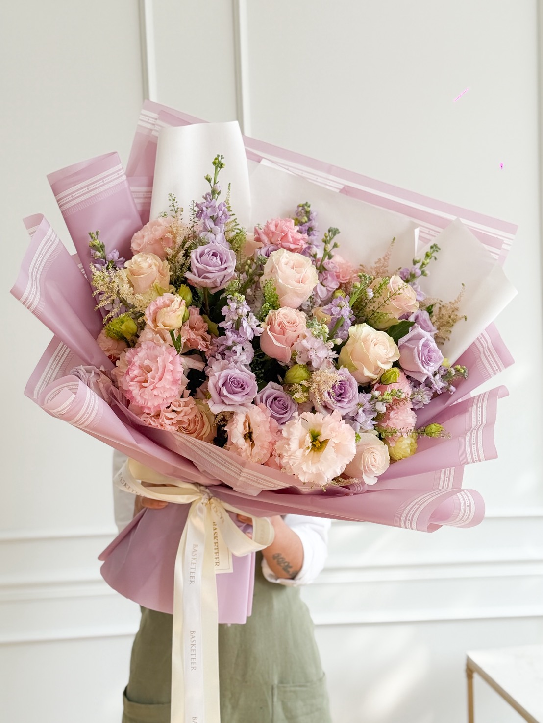 A person holding a Romantic Blossom Bouquet of pink and lavender flowers, wrapped in pastel pink paper and tied with a cream ribbon, stands against a white wall.