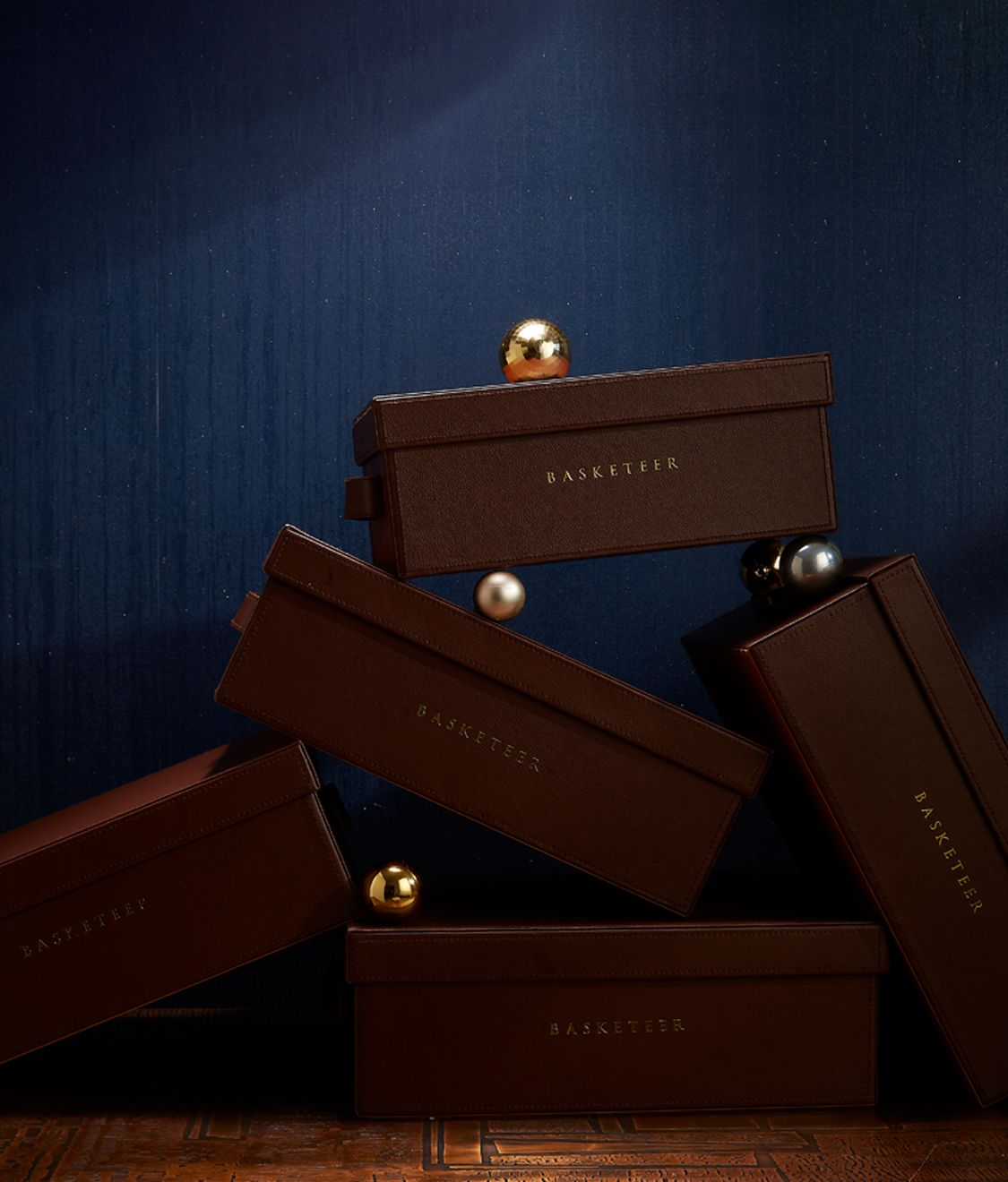 Luxurious dark brown gift boxes embossed with 'Basketeer' in gold, elegantly stacked with metallic decorative spheres