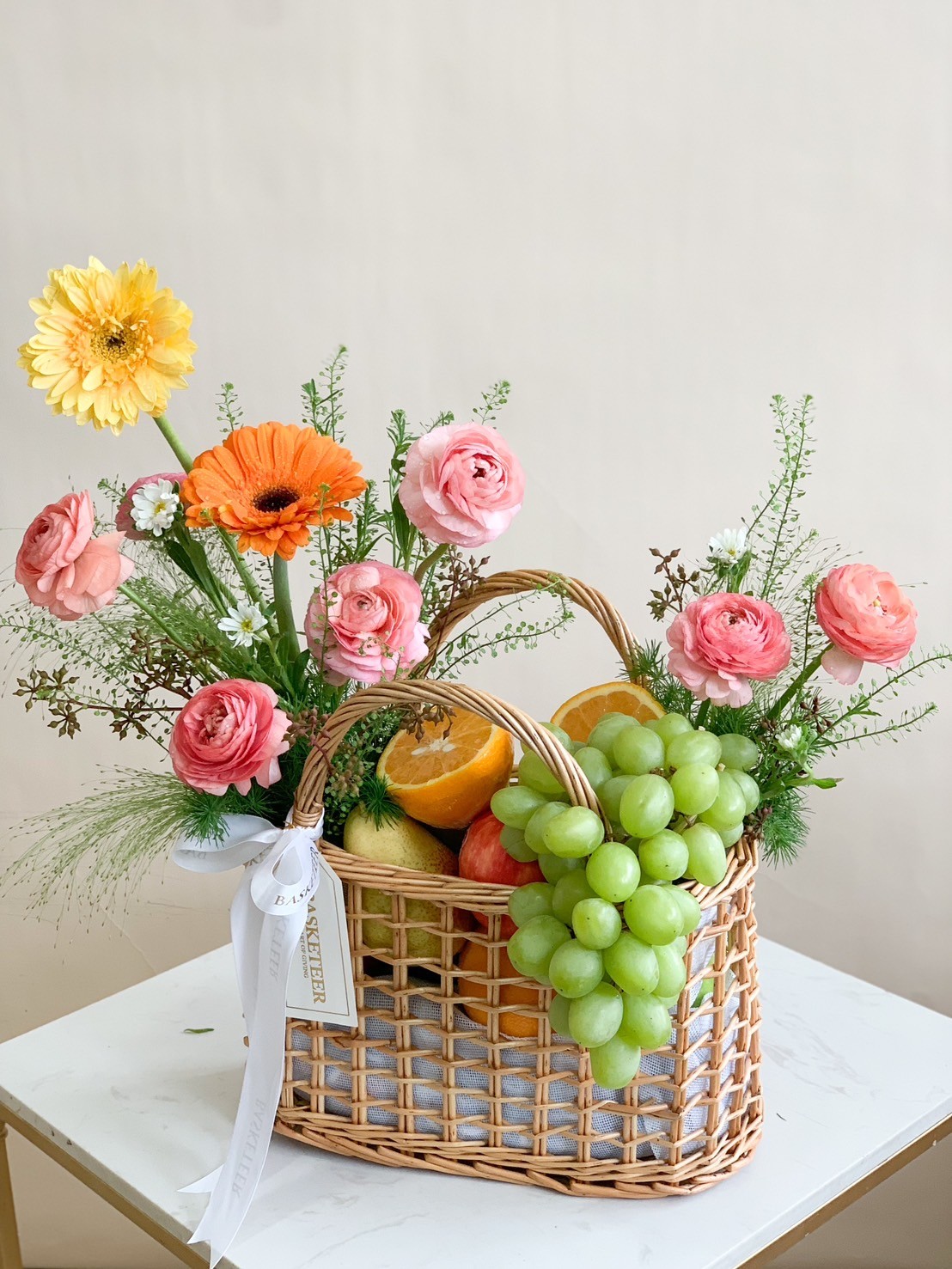 A wicker basket filled with orange and yellow flowers, green grapes, and assorted fruits like green apples, pears, and oranges. Above the basket, more fruits are floating against a neutral background. Some fruits are placed around the basket.