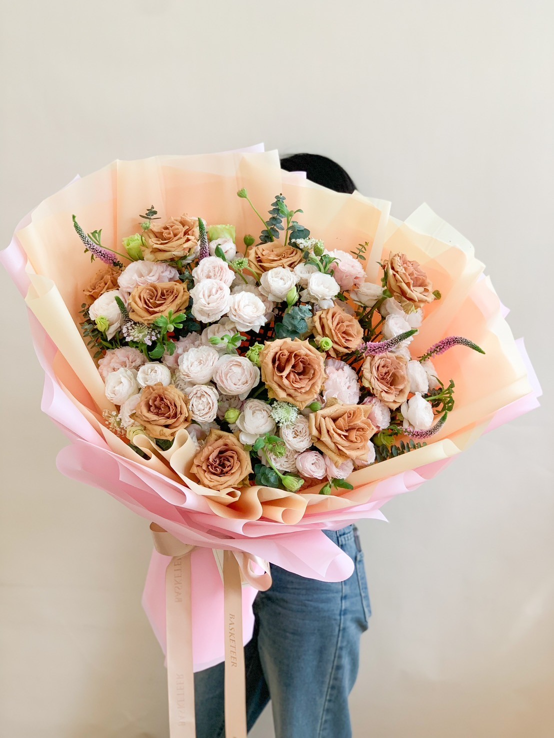 Bouquet of caramel-toned roses, cream blooms, greenery, and wildflowers wrapped in pastel and golden-hued paper, tied with a satin ribbon.