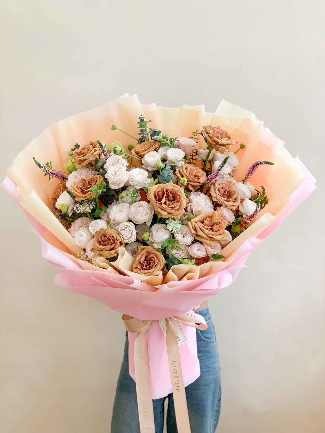 Bouquet of caramel-toned roses, cream blooms, greenery, and wildflowers wrapped in pastel and golden-hued paper, tied with a satin ribbon.